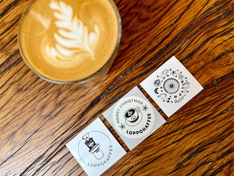Drei runde Aufkleber auf einem Holztisch, daneben steht eine Tasse Flat White Coffee